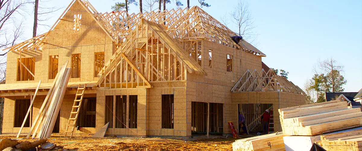 Wooden house under construction with visible framing and roof trusses, surrounded by building materials on a sunny day.